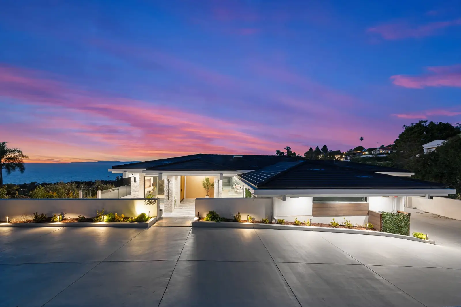 Single-story modern home at twilight with warm landscape lighting and an ocean horizon under a pink sky